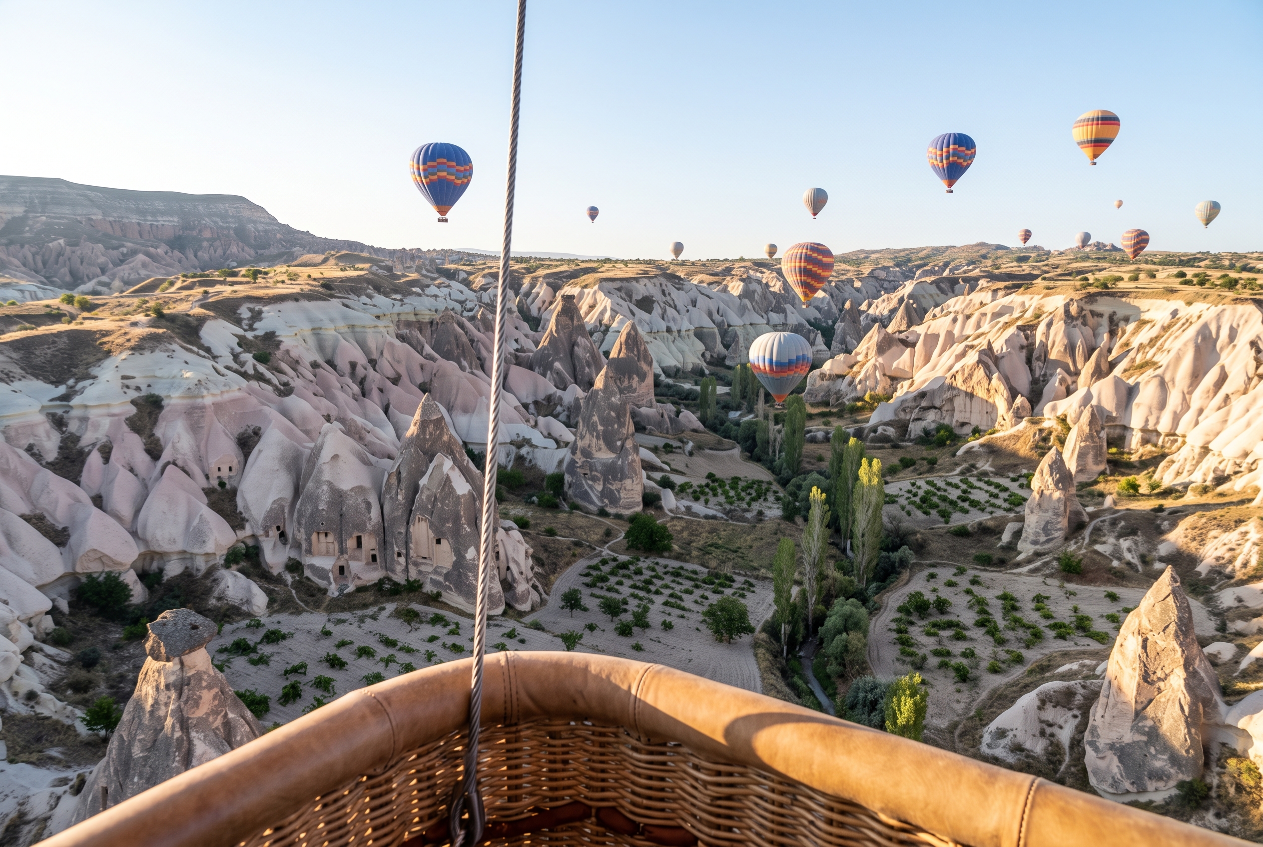 Blick aus einem Ballonkorb über Kappadokien kurz nach Sonnenaufgang: im Vordergrund der Weidenrand und ein Stahlseil, darunter gebänderte Tuffkegel und gehauene Türöffnungen, über dem Tal weitere Ballone in verschiedenen Höhen.